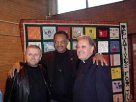 Jesse Jackson in front of the Remembrance Quilt with Holy Cross priests Gary Donnegan and Aidan Troy